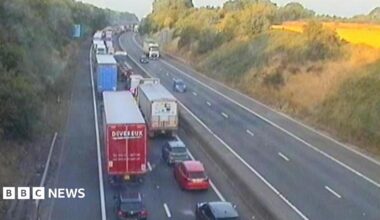A motorway camera image showing a long queue of vehicles on the carriageway to the left, with three vehicles travelling on the right side of the carriageway