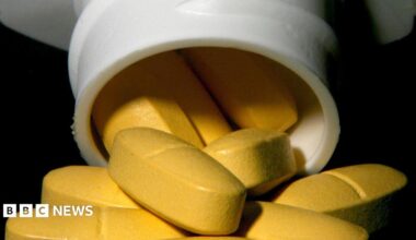 Beatles star Sir Paul McCartney smiles and waves from a car window while holding up a smartphone. Ringo Starr can be seen on the screen, wearing sunglasses. McCartney is dressed in a beige jumper with a light blue shirt collar underneath and several bracelets on his wrist.