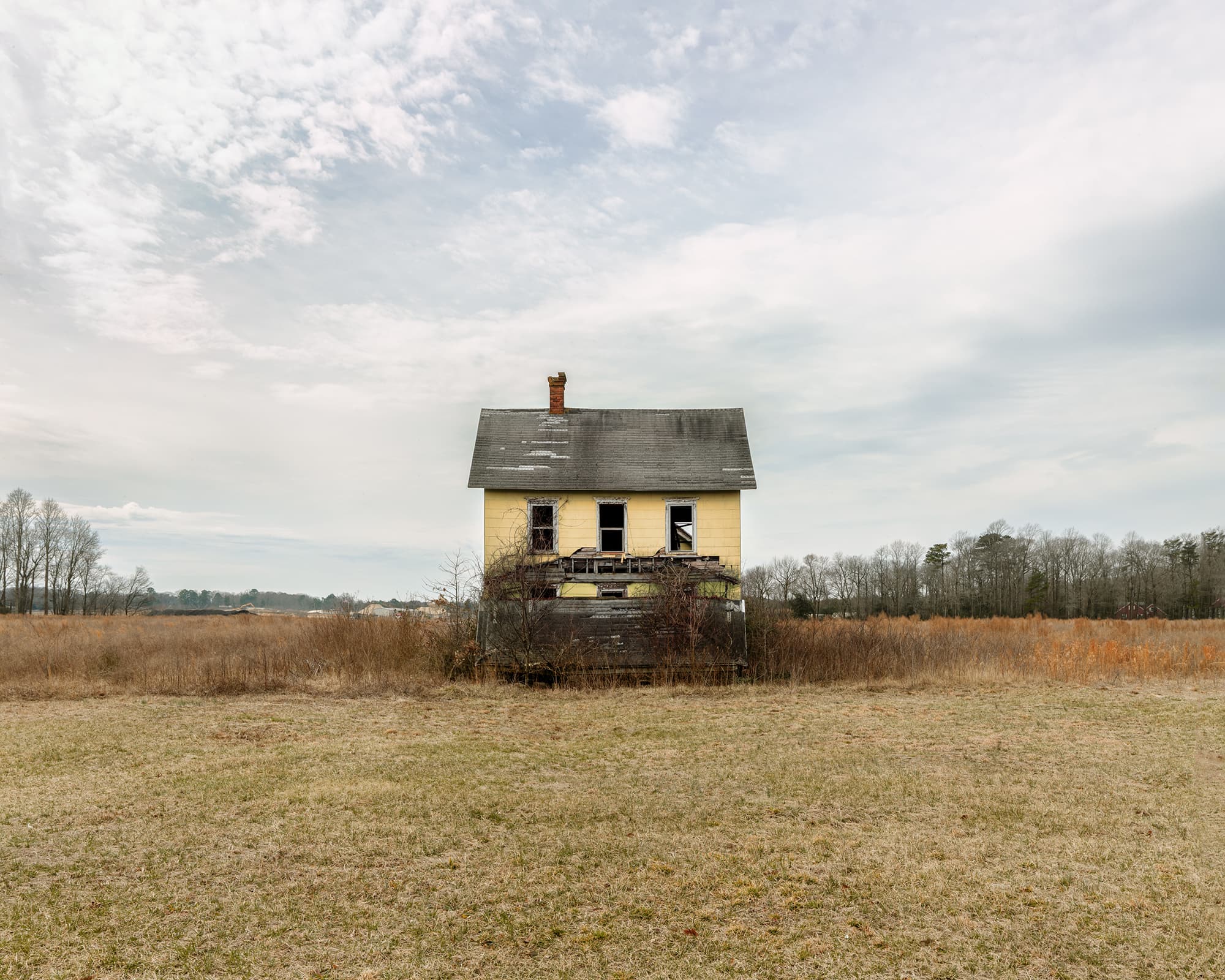 A small, isolated yellow two-story house with a damaged facade sits alone in a vast, flat grassy field stretching toward a distant tree line.