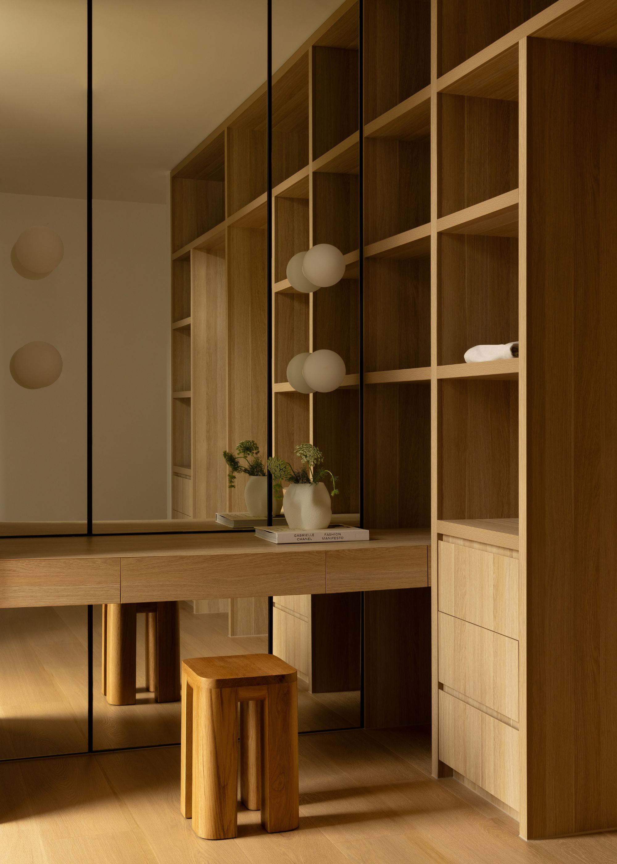 A room with a floating desk, a wood stool, shelves, a mirrored wall and spherical sconces