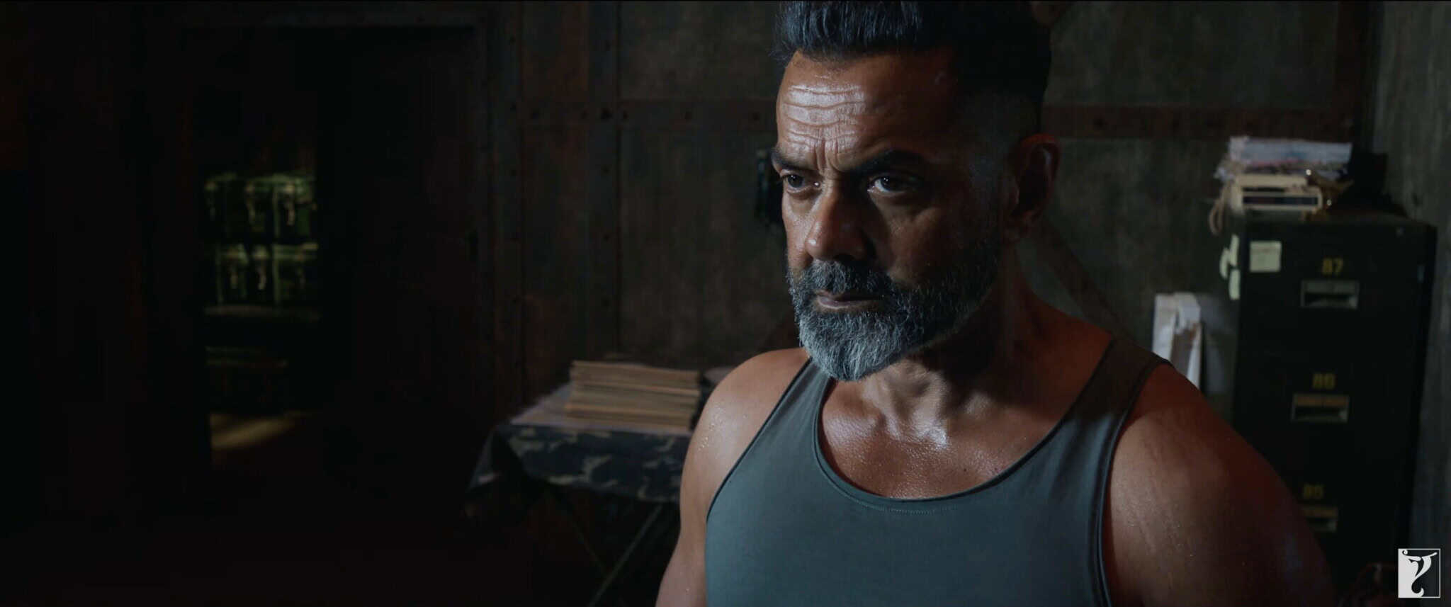 A serious man with a gray beard and mustache, wearing a tank top, stands in a dimly lit room with metal walls, stacks of papers, and a cabinet in the background.