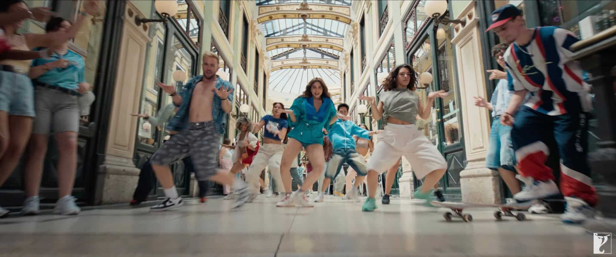A group of people dance energetically in a bright, glass-roofed arcade, with onlookers clapping and watching. Some dancers wear colorful outfits, and one person rides a skateboard in the lively scene.