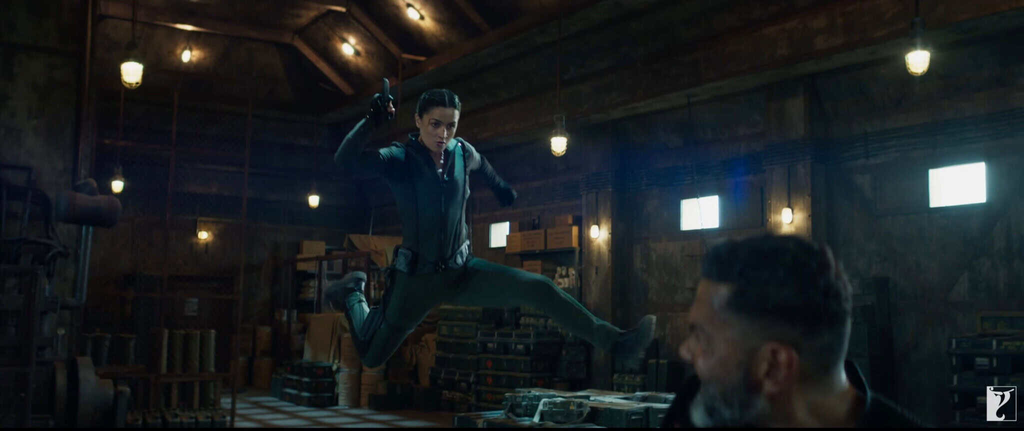 A woman in a dark jumpsuit is mid-air, performing a flying kick towards a man in a dimly lit warehouse filled with crates and metal shelves. Bright lights hang from the wooden ceiling above.