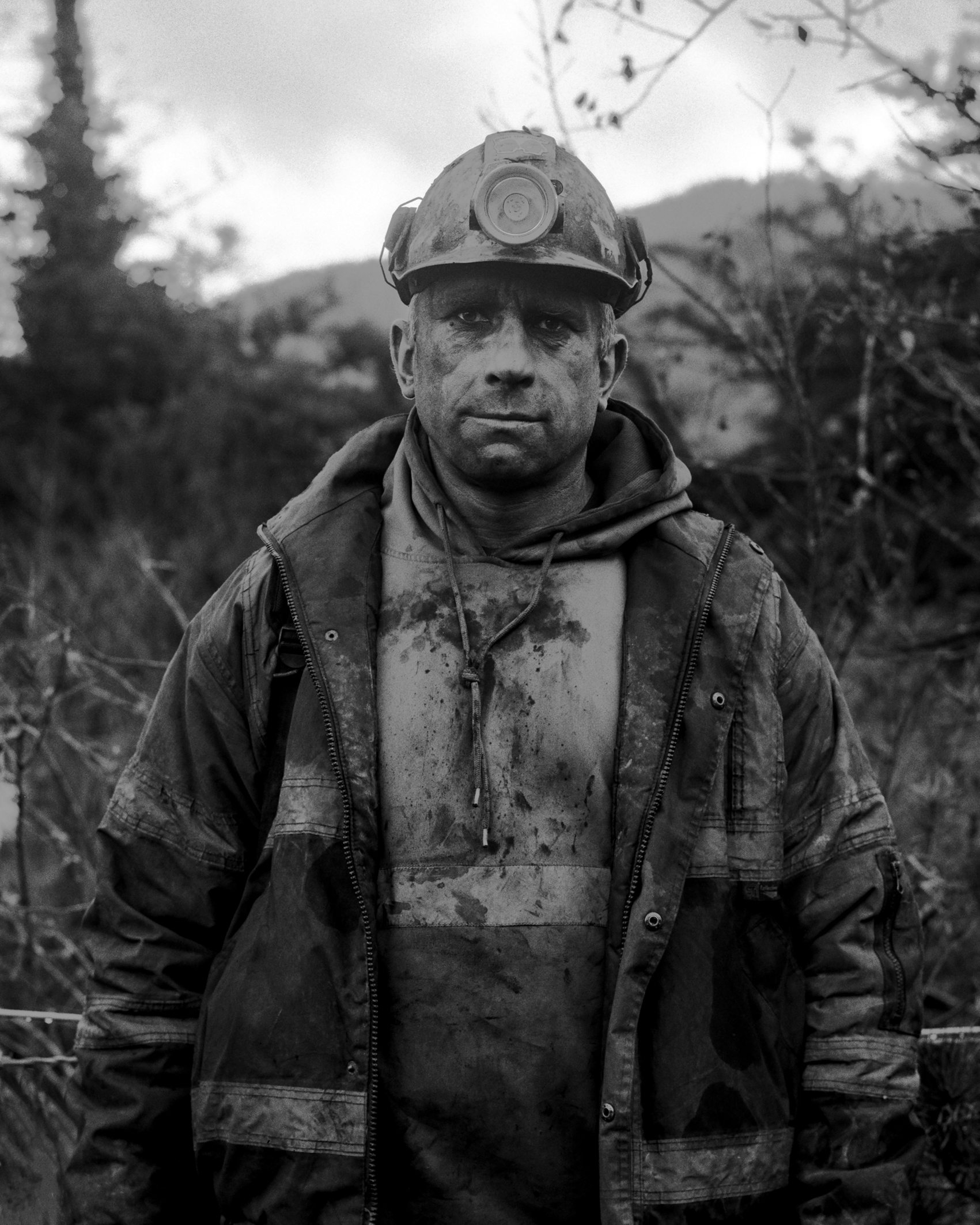 Black and white photo of a miner wearing a helmet and dirty work clothes, standing outdoors with trees and hills in the background