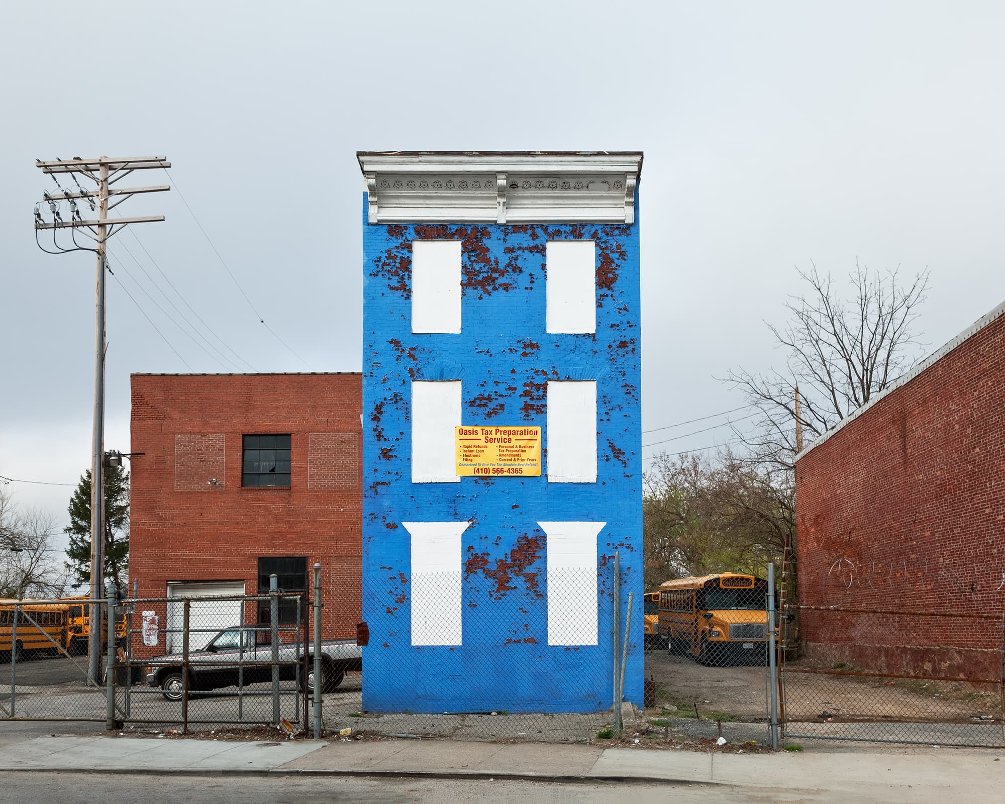 A standalone three-story building painted bright blue features blocked-out white windows and an "Oasis Tax Preparation Service" sign, situated next to an empty lot filled with yellow school buses.