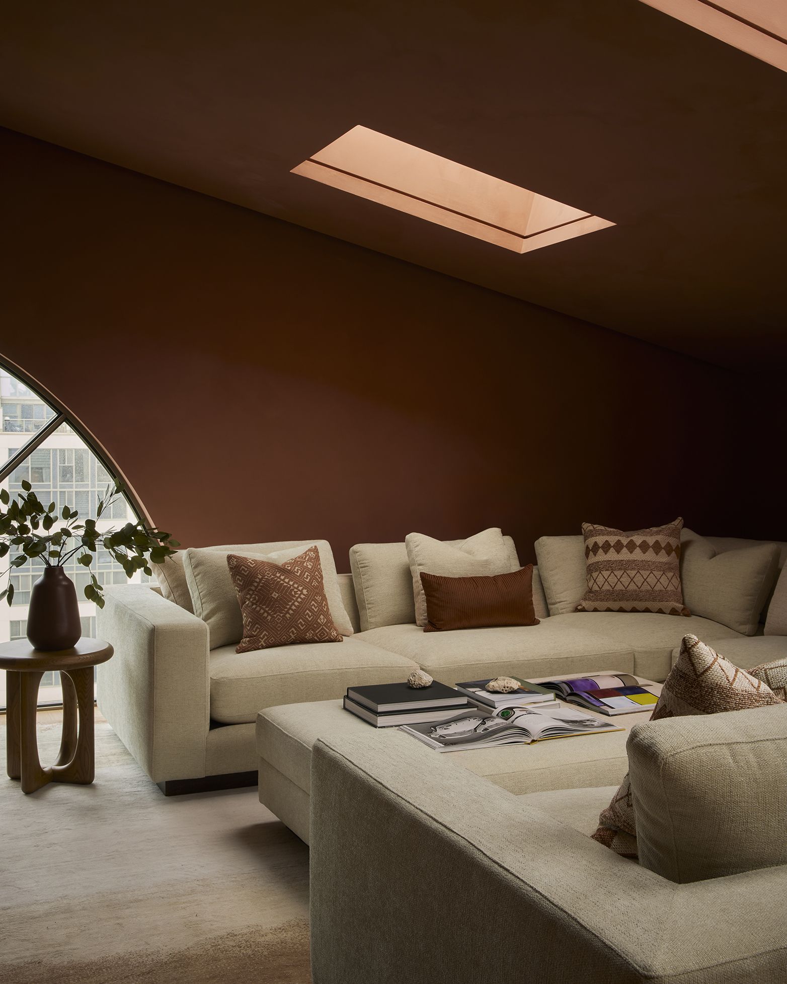 A terracotta-painted living room with two facing beige sofas, decorated with terracotta cushions, wooden coffee tables topped with vases, and skylights.