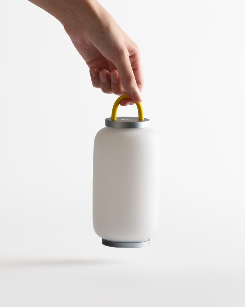 A hand holding a modern, cylindrical white lantern with a yellow handle from DailyObjects stands out against a plain white background.
