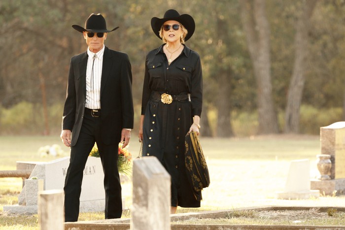 An older man and woman dressed in black western-style clothing walk through a cemetery in daylight. Both wear black cowboy hats and sunglasses, with the woman also wearing a black dress with gold buttons and a large gold belt buckle. Gravestones surround them as they walk side by side with solemn, composed expressions.