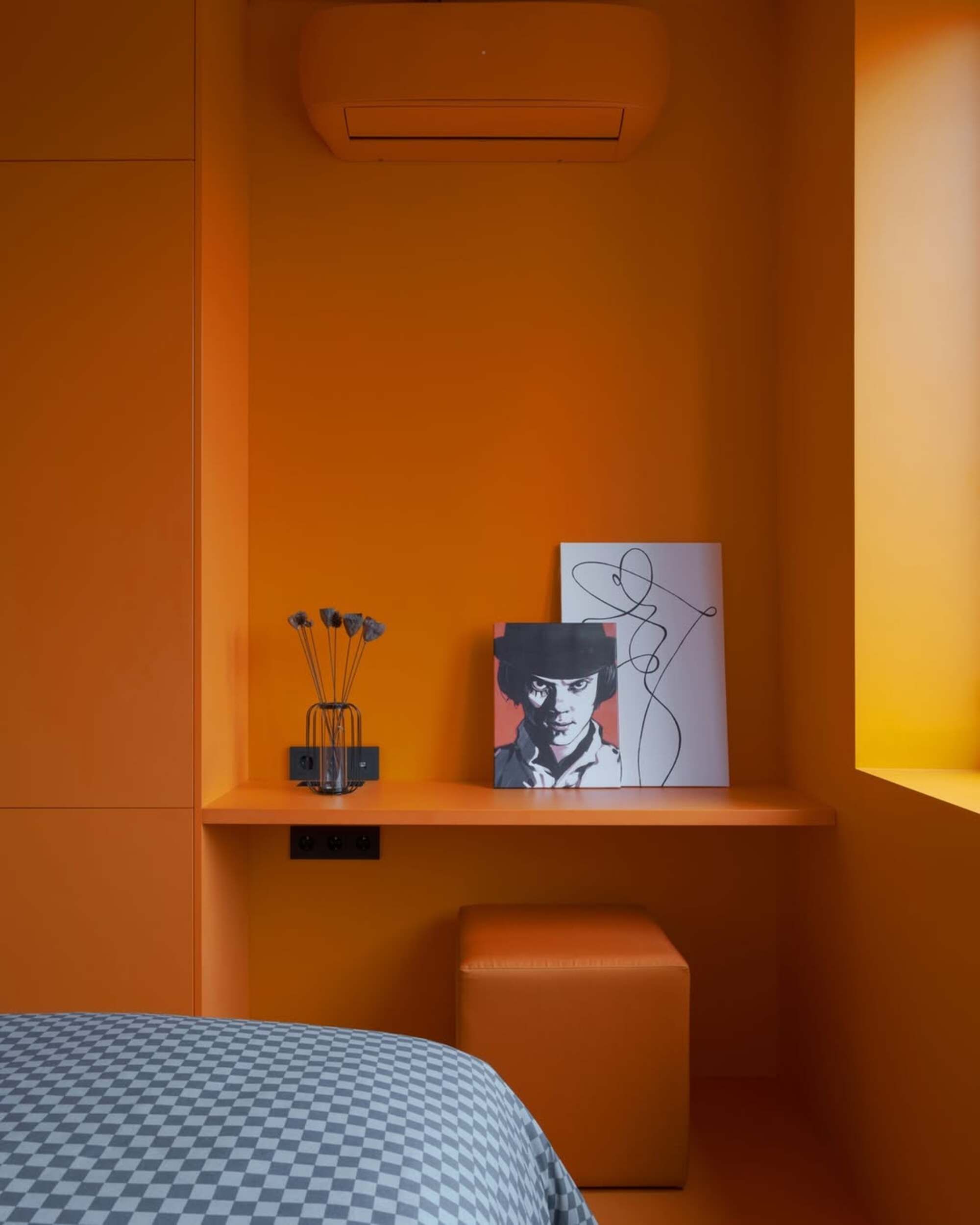Image of a modern room drenched in a bright mango orange color. There is a desk area with a couple of paintings on a shelf and an orange pouf. The corner of a blue bed is in view.
