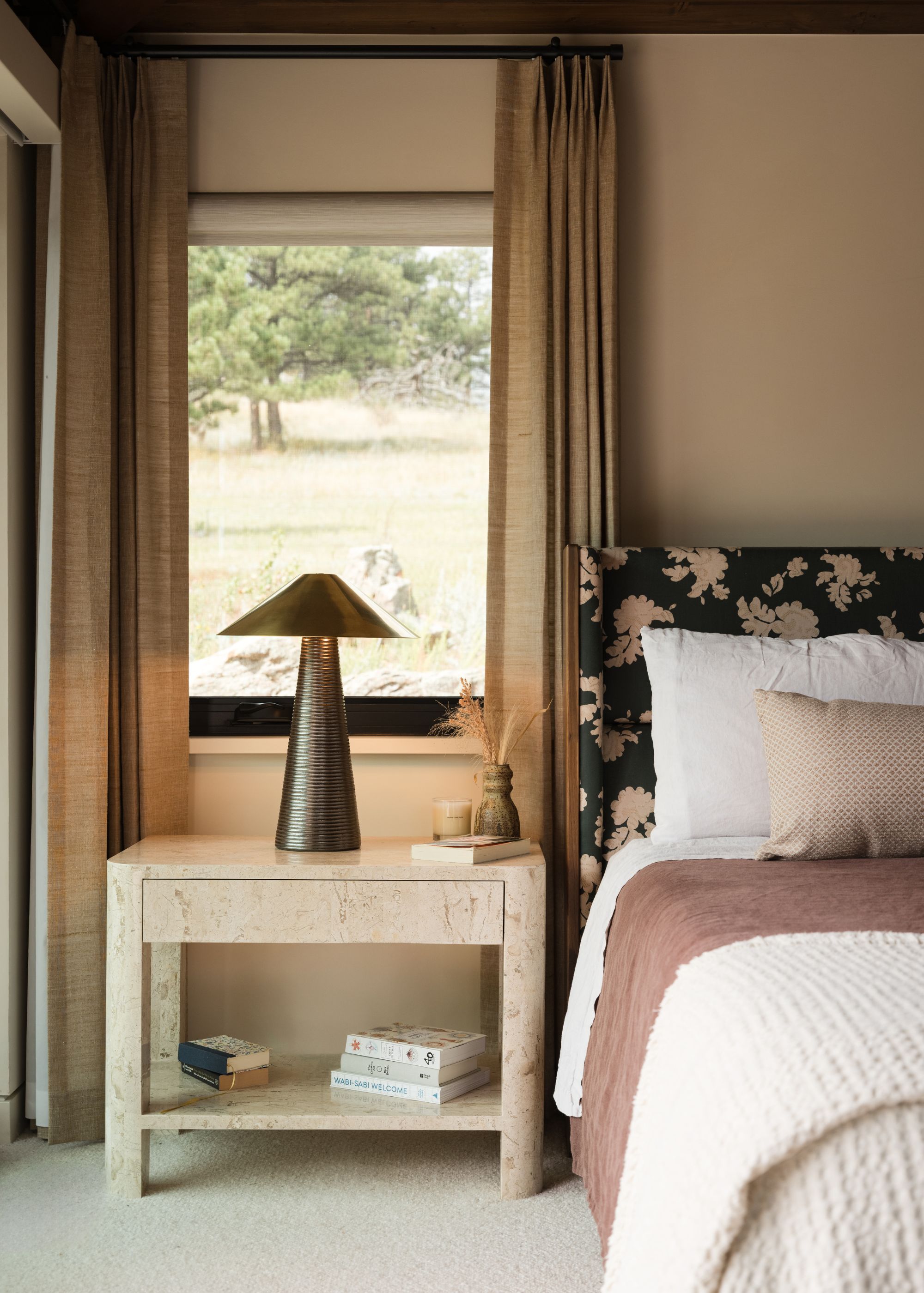 A bedroom with linen curtains, a metallic table lamp on a stone nightstand beside a patterned headboard and a white and clay bedscape