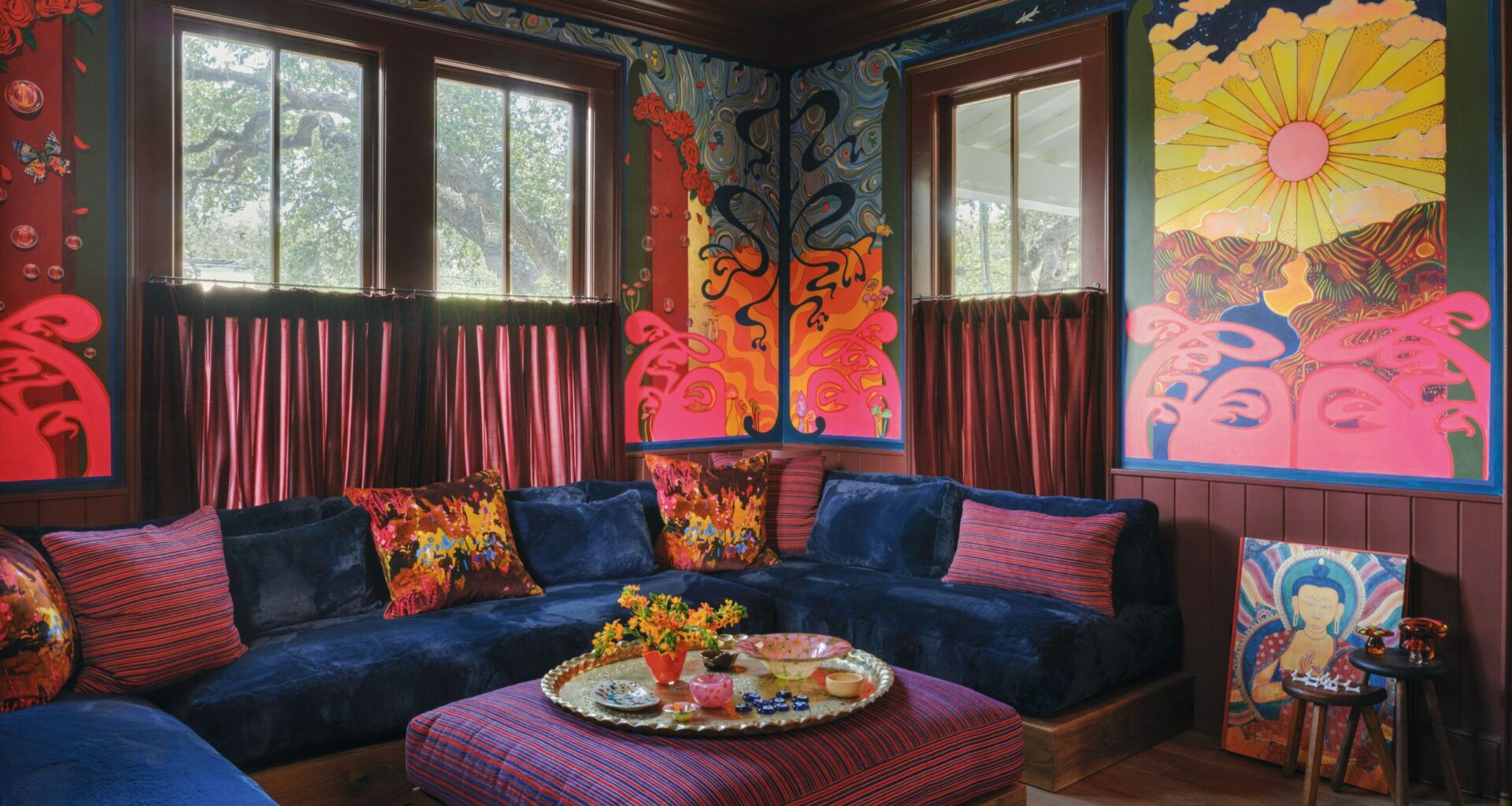 A vibrant, colorful living room corner with a built-in sofa with plush blue velvet seating and colorful scatter cushions, a purple striped pouf in the center with a round tray with plates and decor