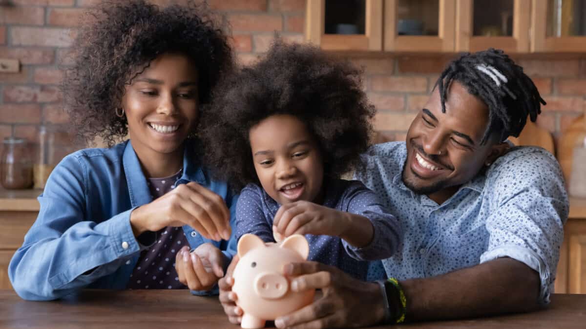 Young girl starting investing by putting a coin in a piggybank while surrounded by her parents.