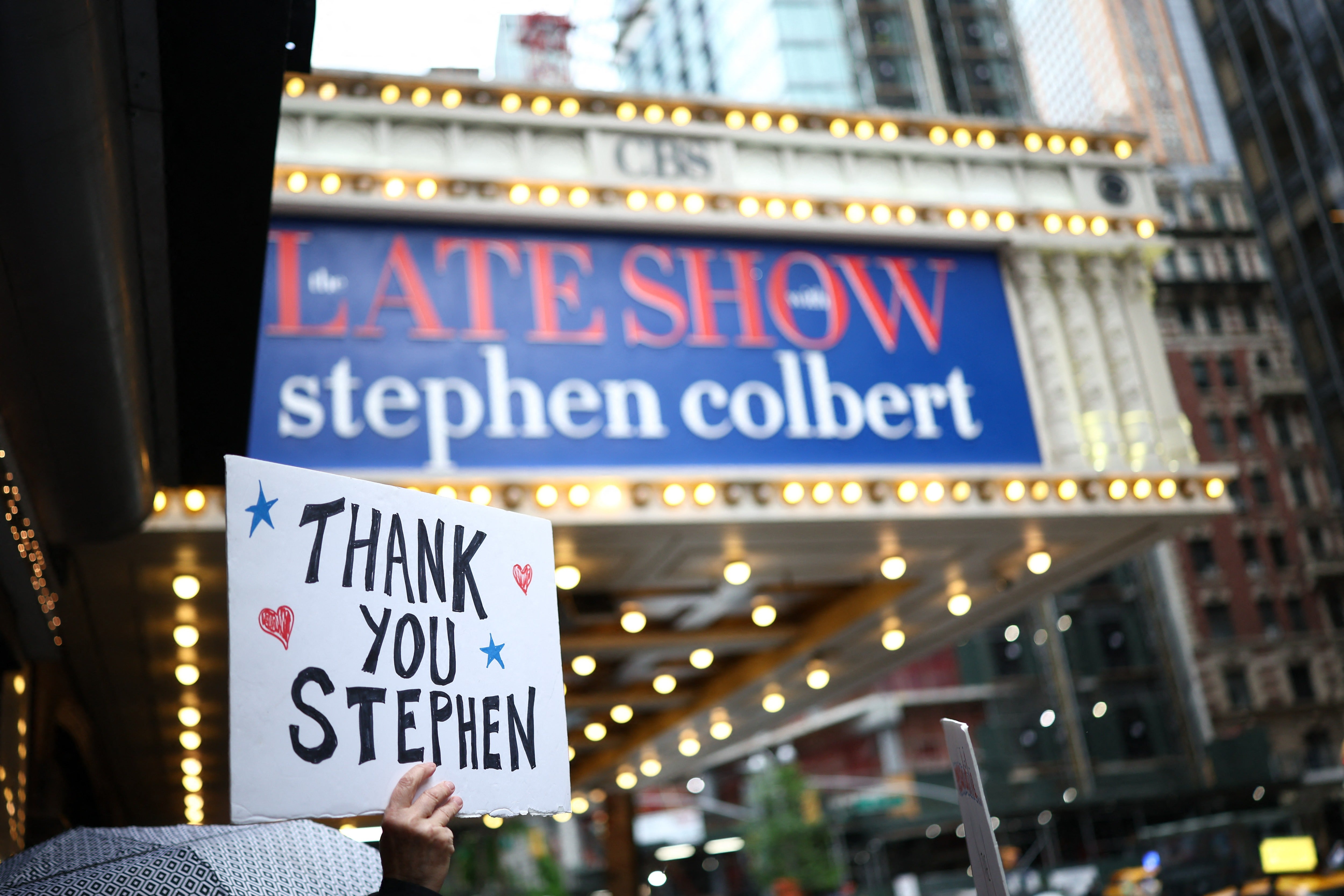 People hold signs supporting Stephen Colbert outside the Ed Sullivan Theater ahead of the taping of the final episode of the The Late Show on May 21, 2026