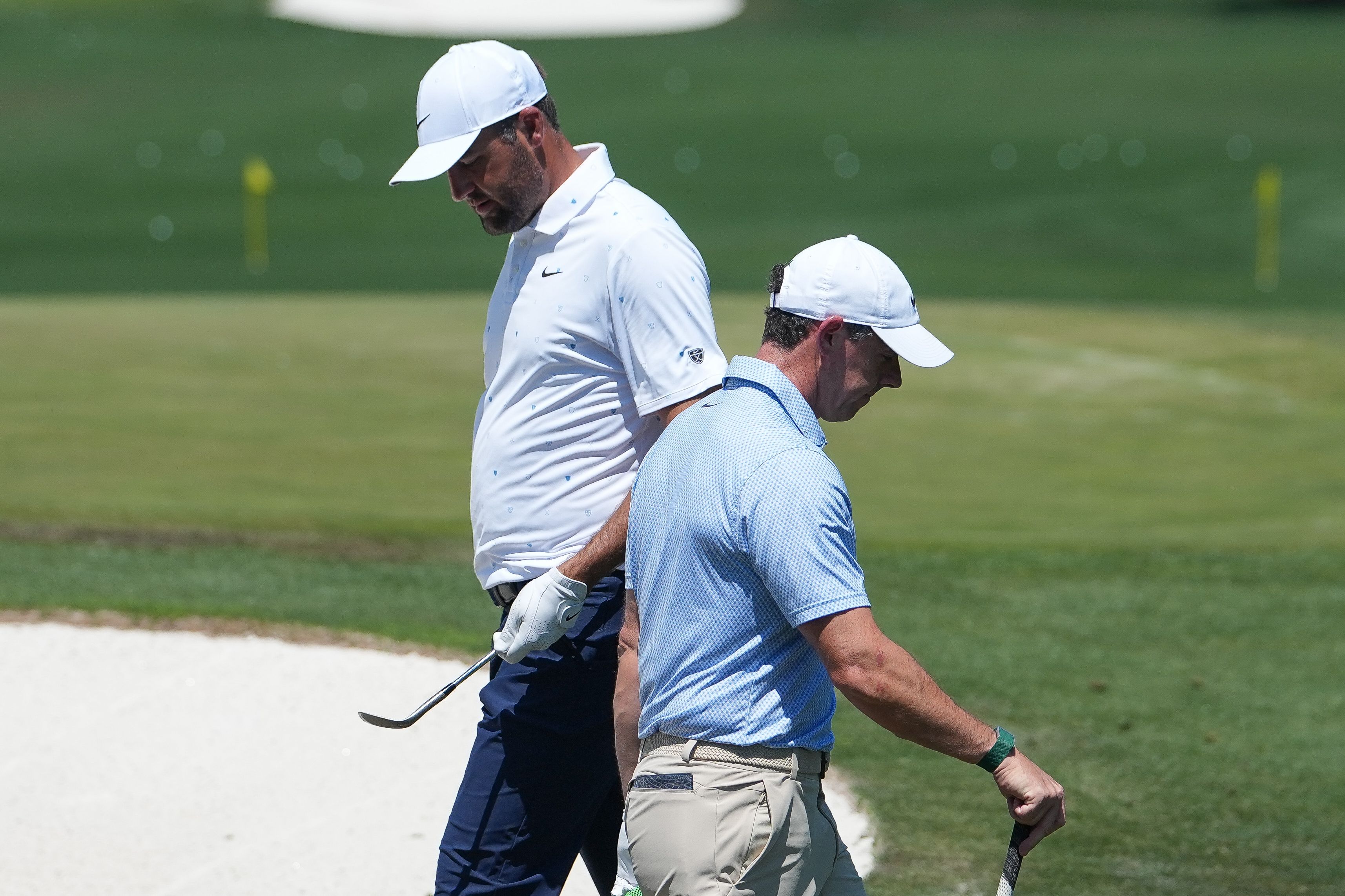 Two golfers in white and blue shirts stand on a green course near a sand trap.