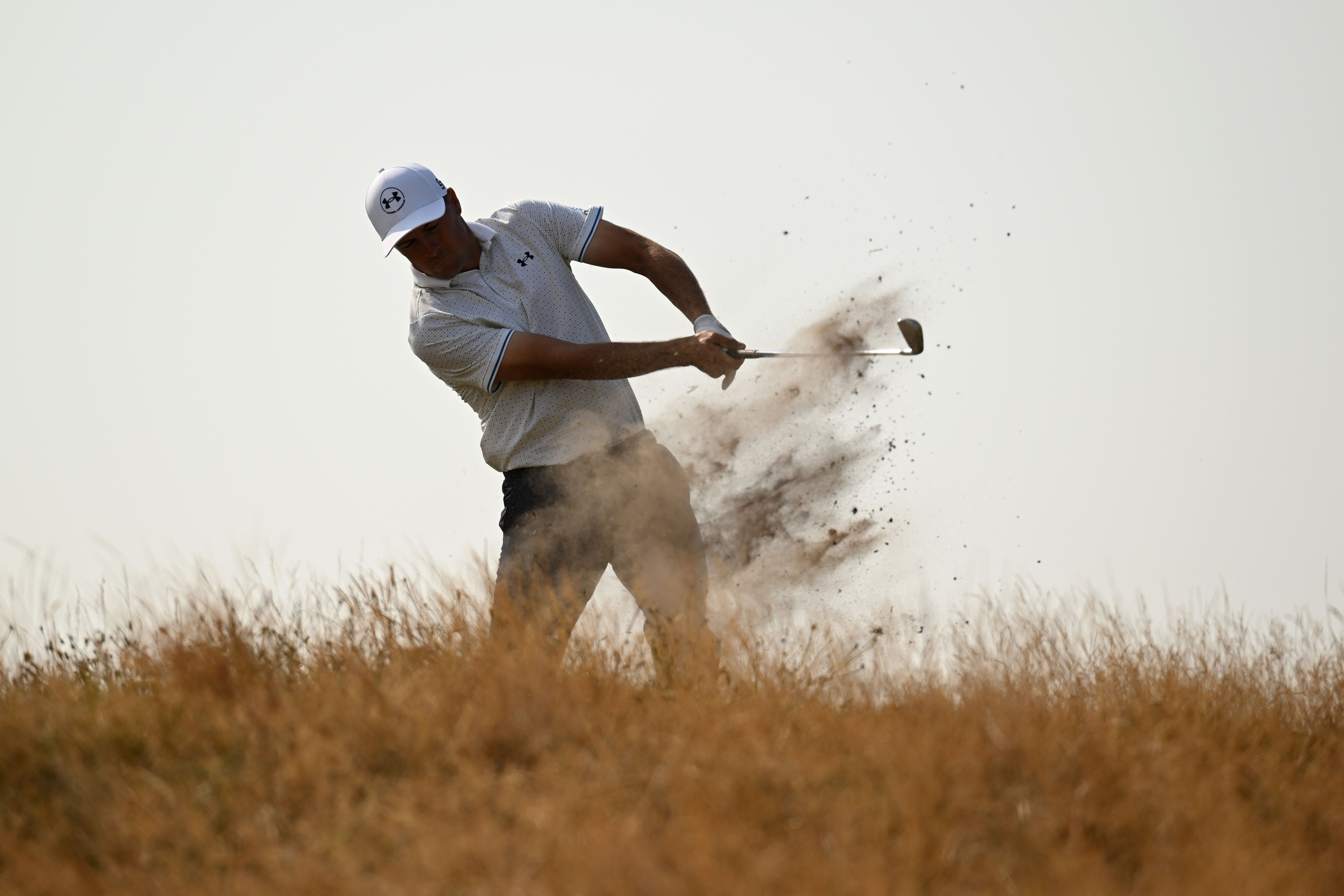 Male golfer powerfully hits golf ball, sending sand flying from a bunker.