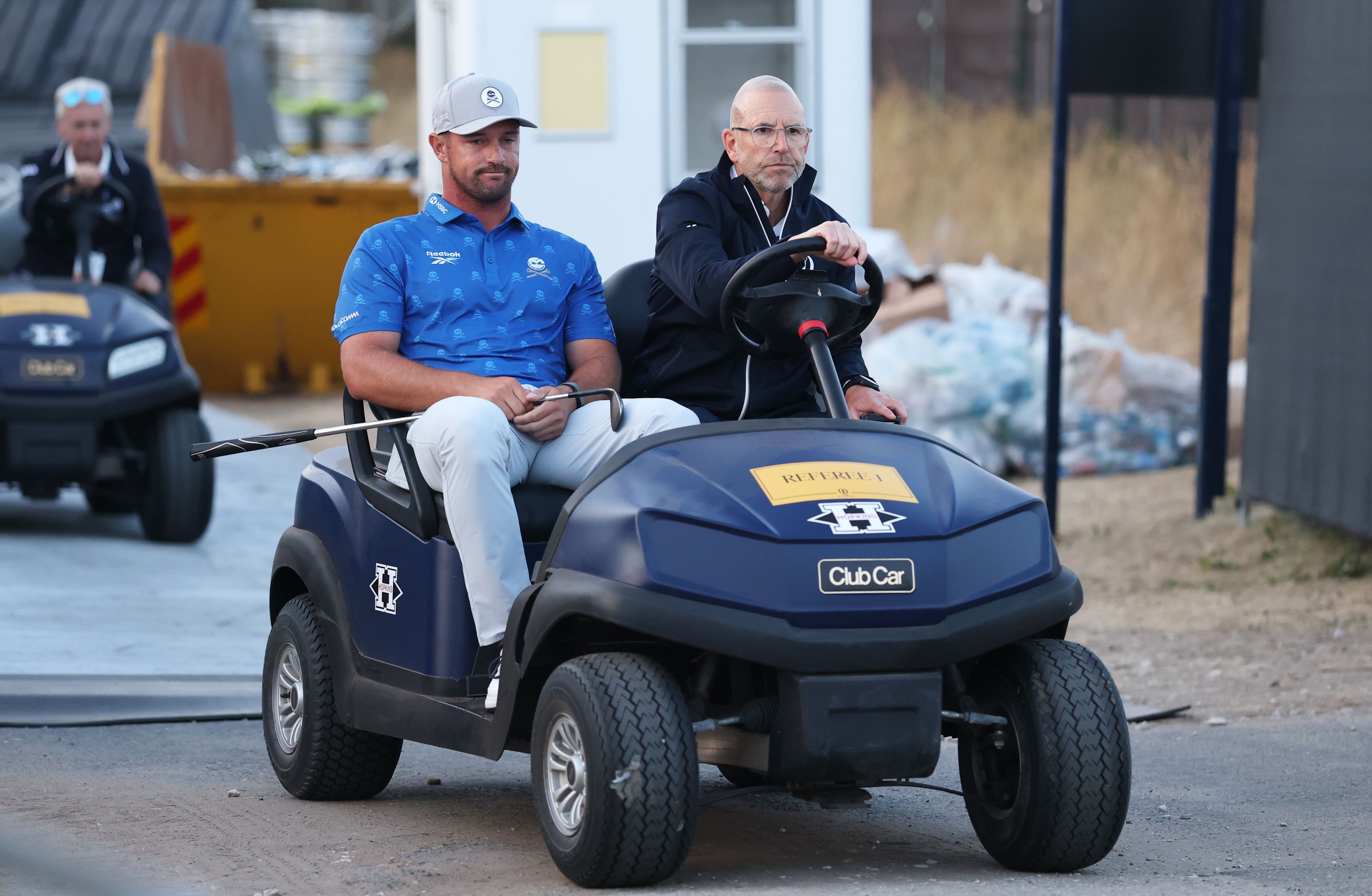 Two men ride in a blue golf cart; one wears a blue polo and cap.