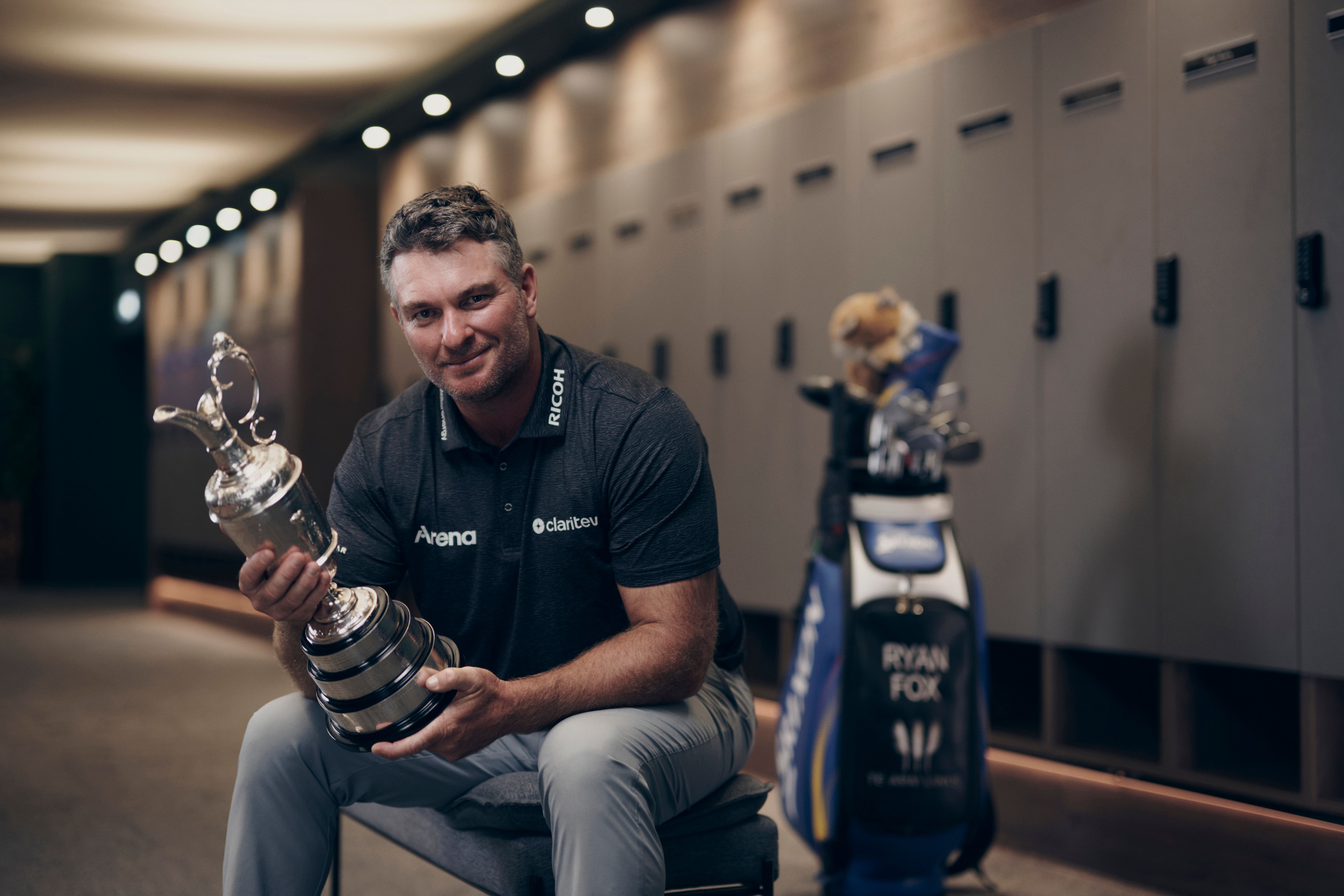 Golfer Bryson DeChambeau smiles with a silver trophy and golf bag in a locker room.