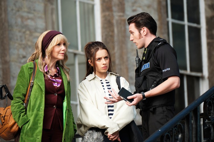 The Hairdresser Mysteries Three people stand outside a stone building in conversation. A police officer in uniform gestures while speaking to two women, one wearing a bright green coat and headband and the other dressed in a light-coloured jacket over a striped top. The younger woman looks concerned as she listens, while the officer appears to be explaining something. The scene takes place on outdoor steps beside large windows, suggesting an ongoing investigation or official inquiry.