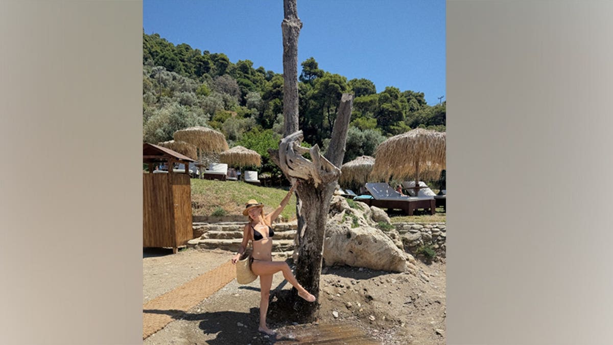 Kate Hudson smiles while leaning against a large tree stump in a black bikini and straw hat during a beach day in Greece.
