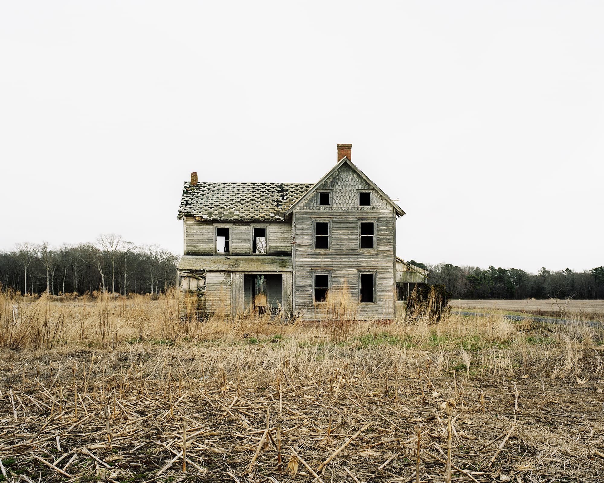 A weathered, gray wooden two-story farmhouse stands derelict and decaying in the middle of a barren, harvested cornfield under an overcast sky.