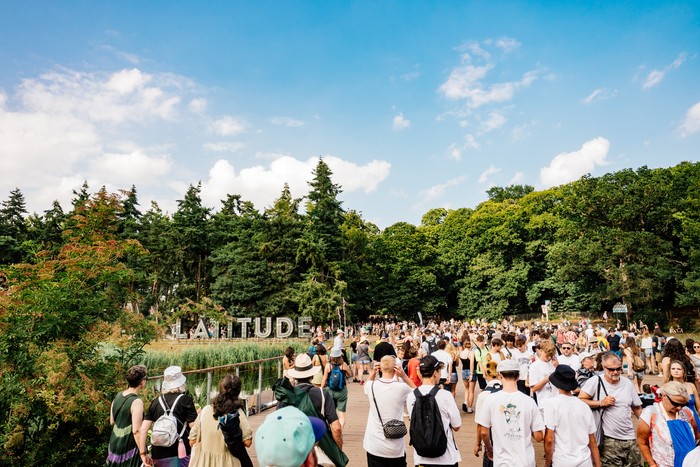Latitude Festival 2025 Crowds walk across a wooden bridge at Latitude Festival, with the large “Latitude” sign, green woodland, and blue summer sky in the background.