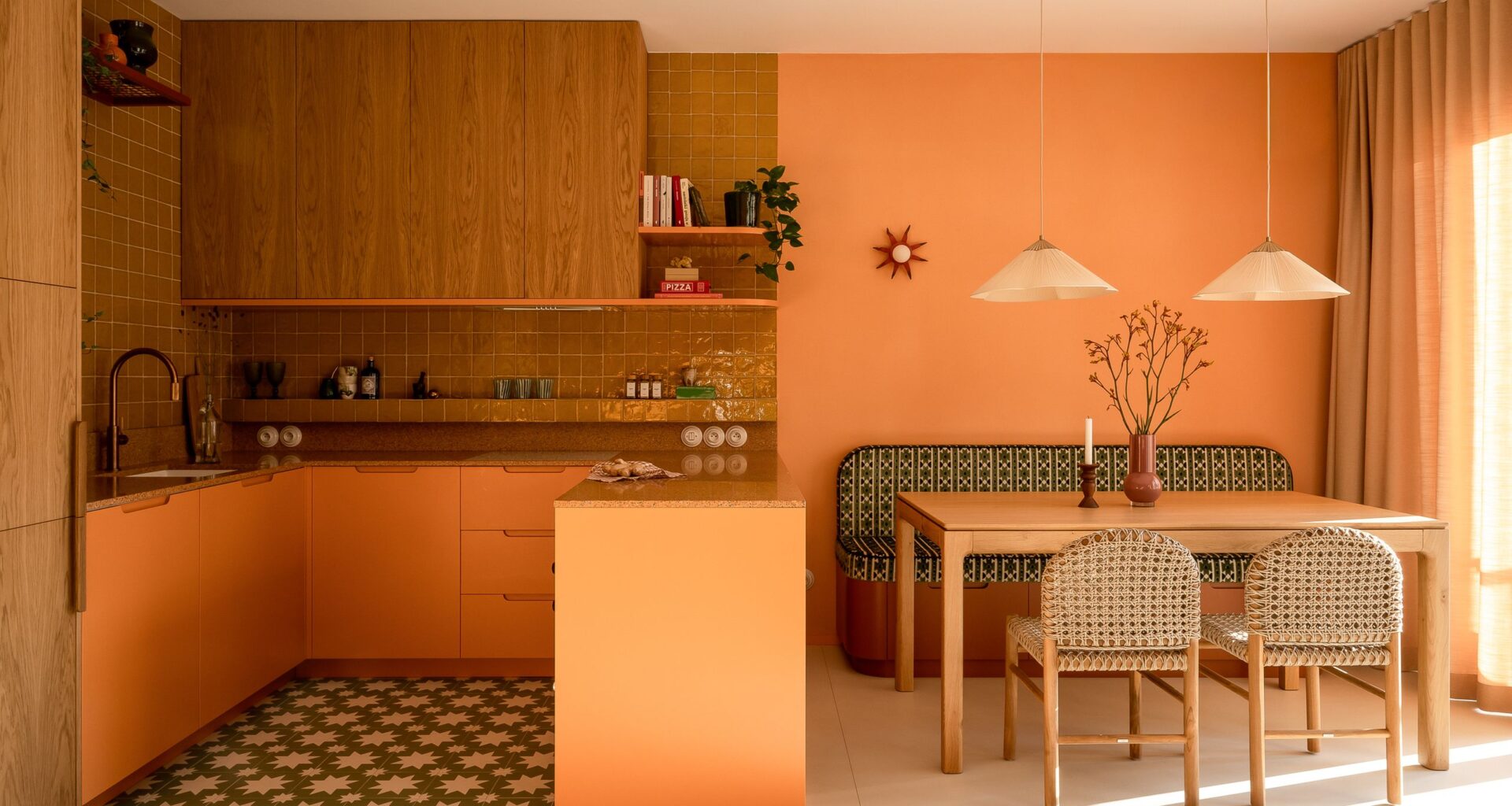 Warm-toned open-plan kitchen and dining room with orange cabinetry, oak cabinets, patterned floor tiles, woven dining chairs, and soft pendant lights