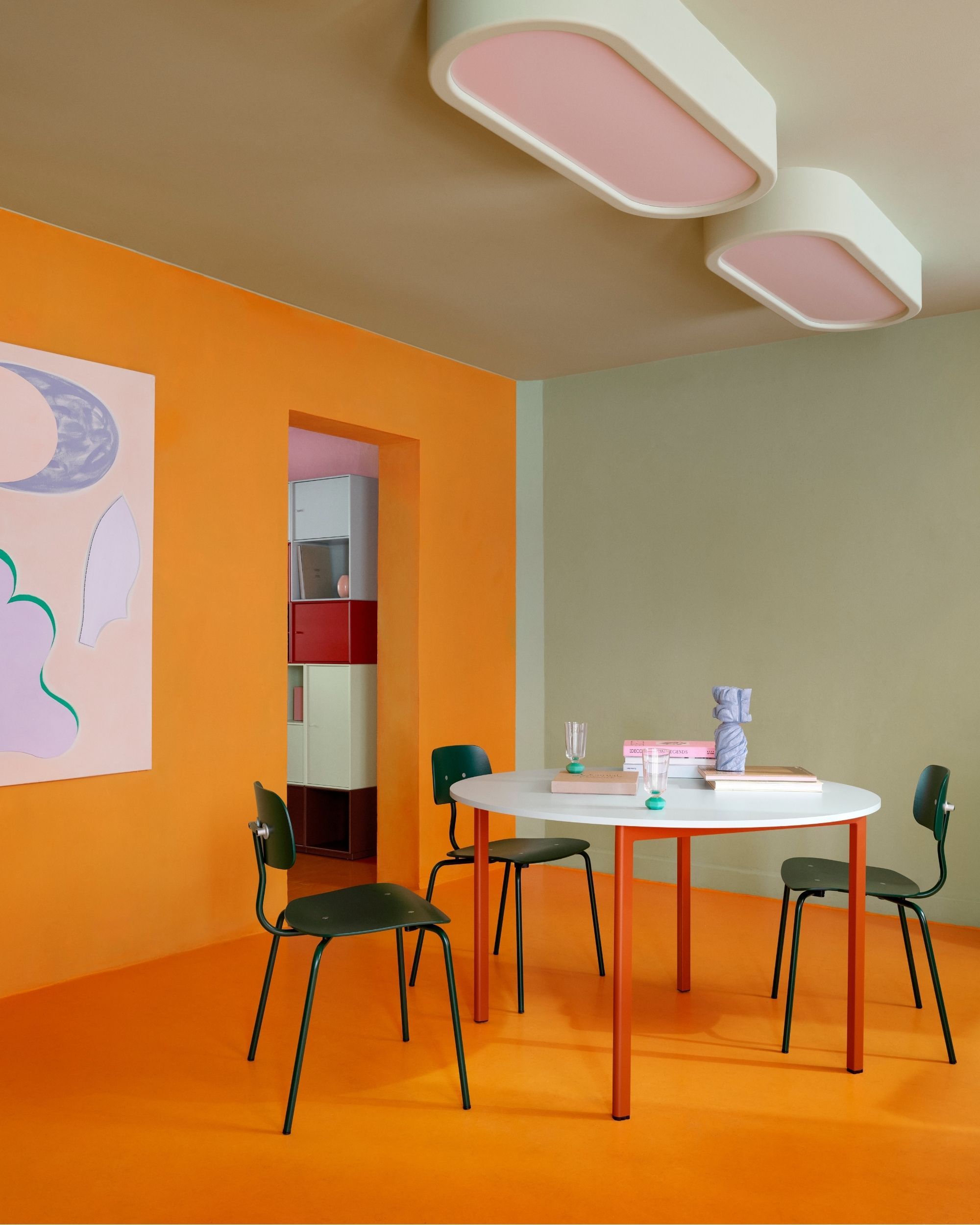 A modern dining room with color-blocked walls. The floor and back wall are orange, the side wall is a light mint green, and the ceiling is cream. There is a red round table with dark green chairs.