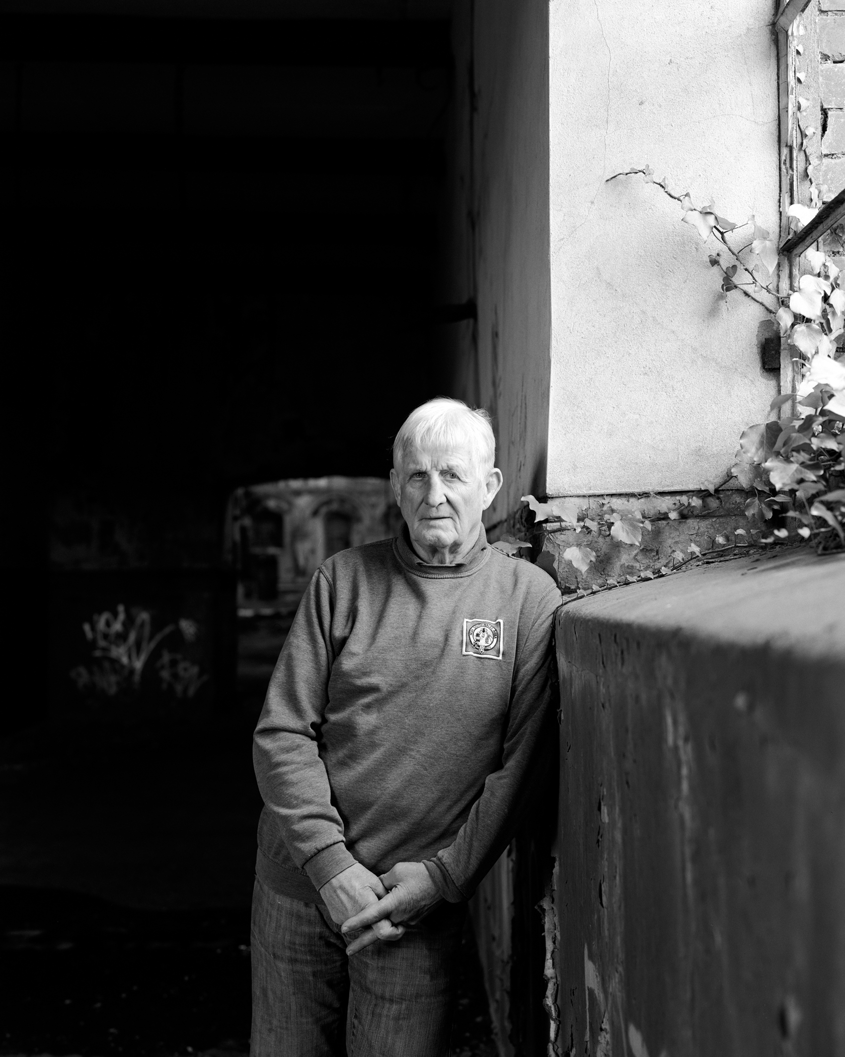 Black and white photo of an elderly man leaning against a cracked wall near a window with vines, in a dimly lit, worn indoor space