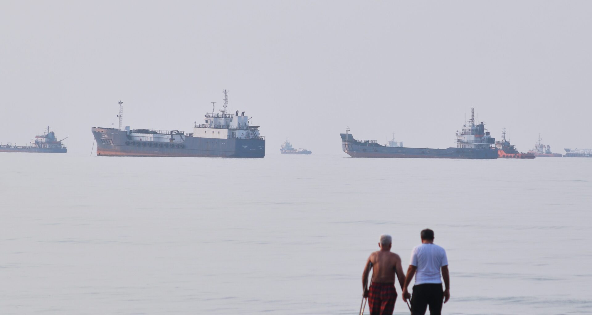 Two men stand in the ocean with multiple oil tankers and cargo ships in the hazy distance.