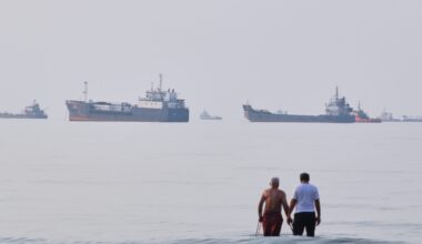 Two men stand in the ocean with multiple oil tankers and cargo ships in the hazy distance.
