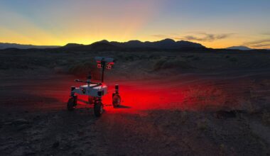 A small rover with lights glowing red underneath stands on rugged terrain at dusk, with mountains silhouetted against a colorful sunset sky in the background.