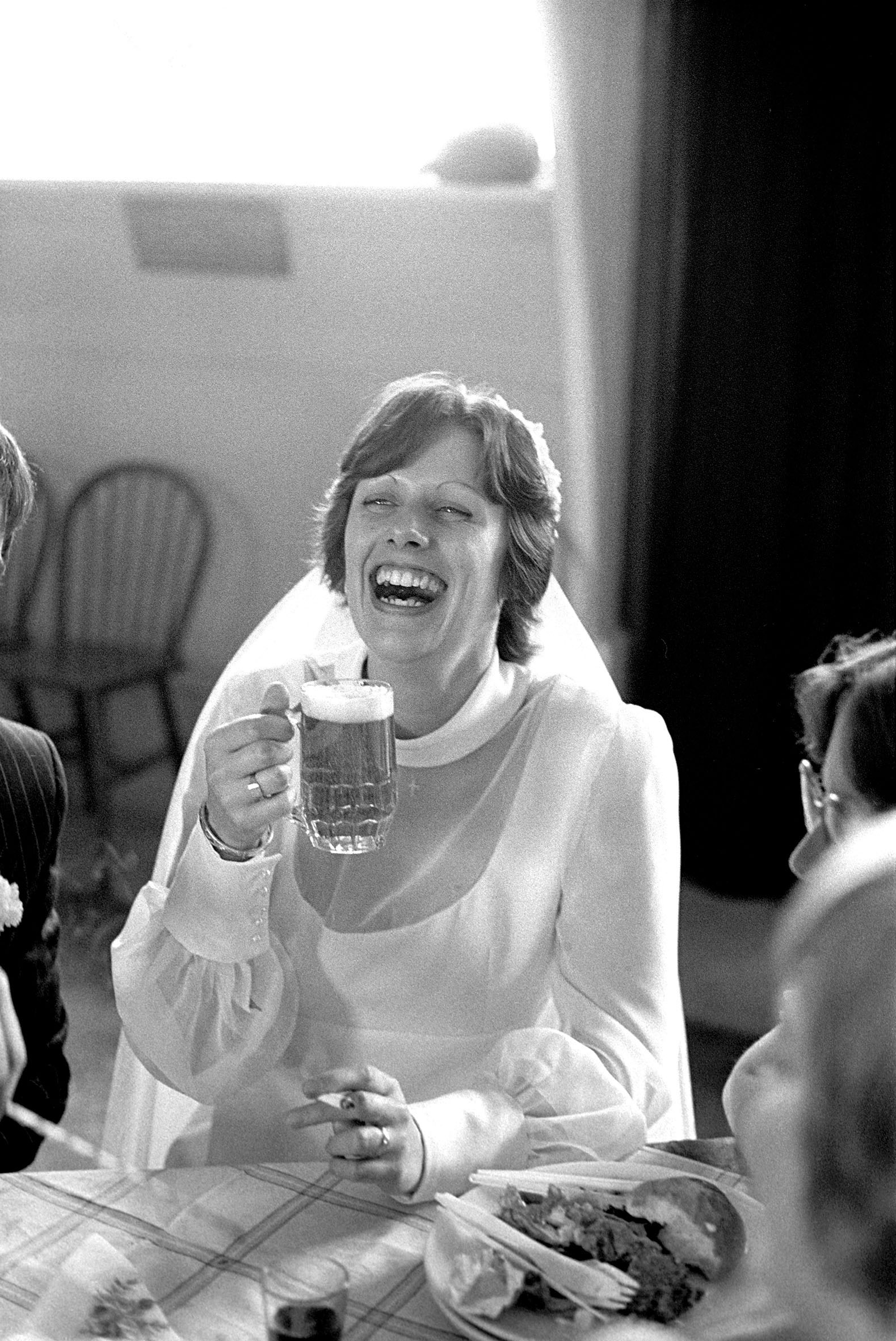 A bride in a white wedding gown and veil laughs heartily while holding a glass mug filled with beer at a reception table.