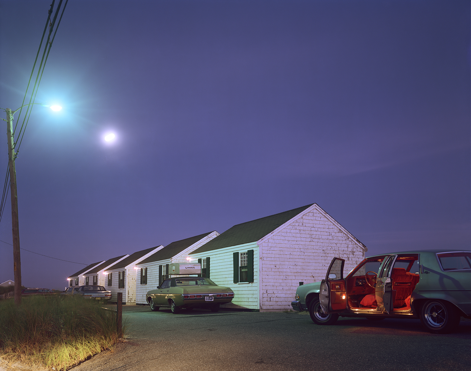 A row of white clapboard cottages glows under a purple night sky, with a green sedan's doors open to reveal its red interior, lit by a streetlamp and the moon above.