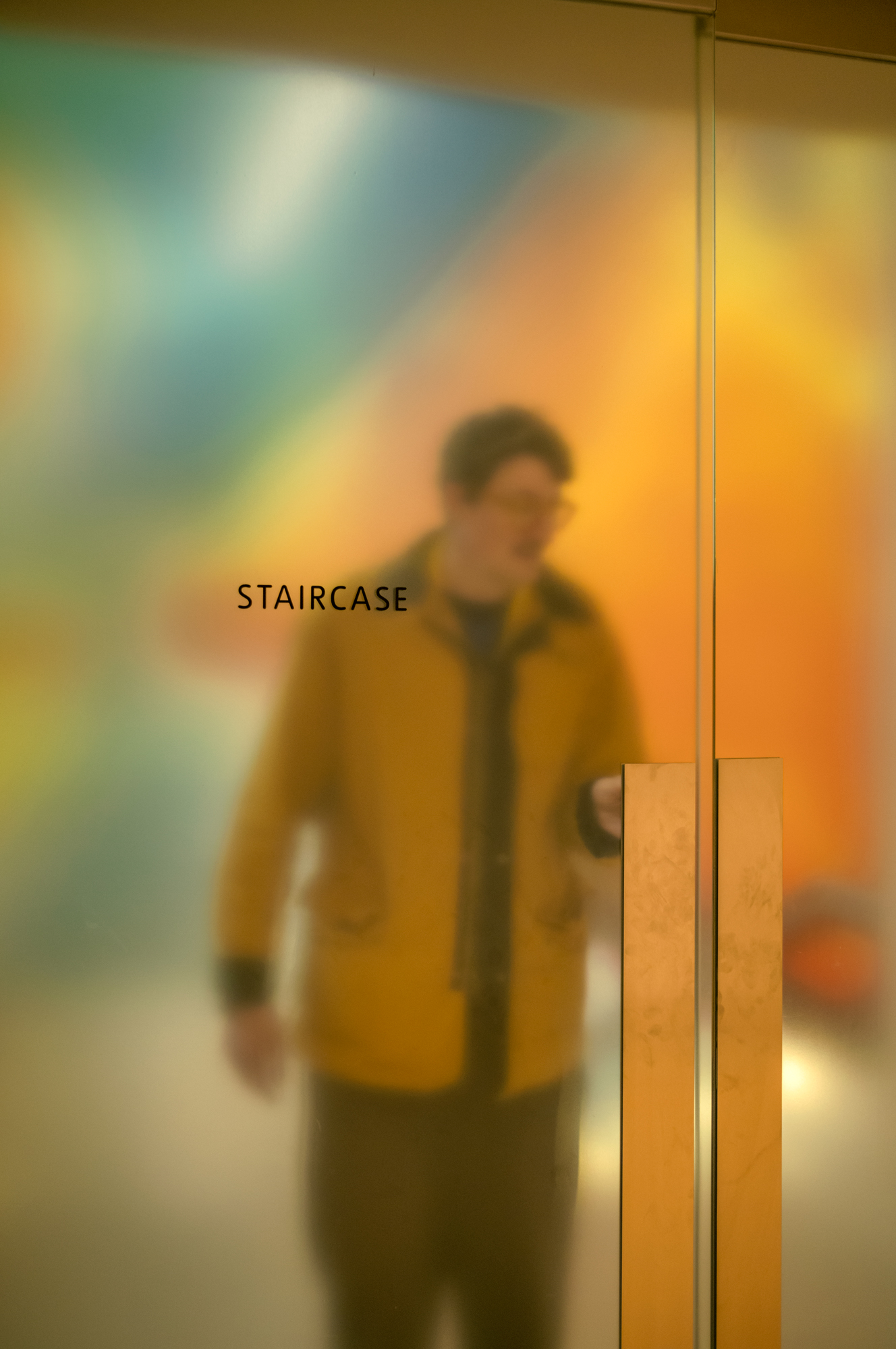 A blurred figure in a mustard-yellow jacket is seen through a glass door labelled "Staircase," reflecting soft washes of teal, orange and yellow light.