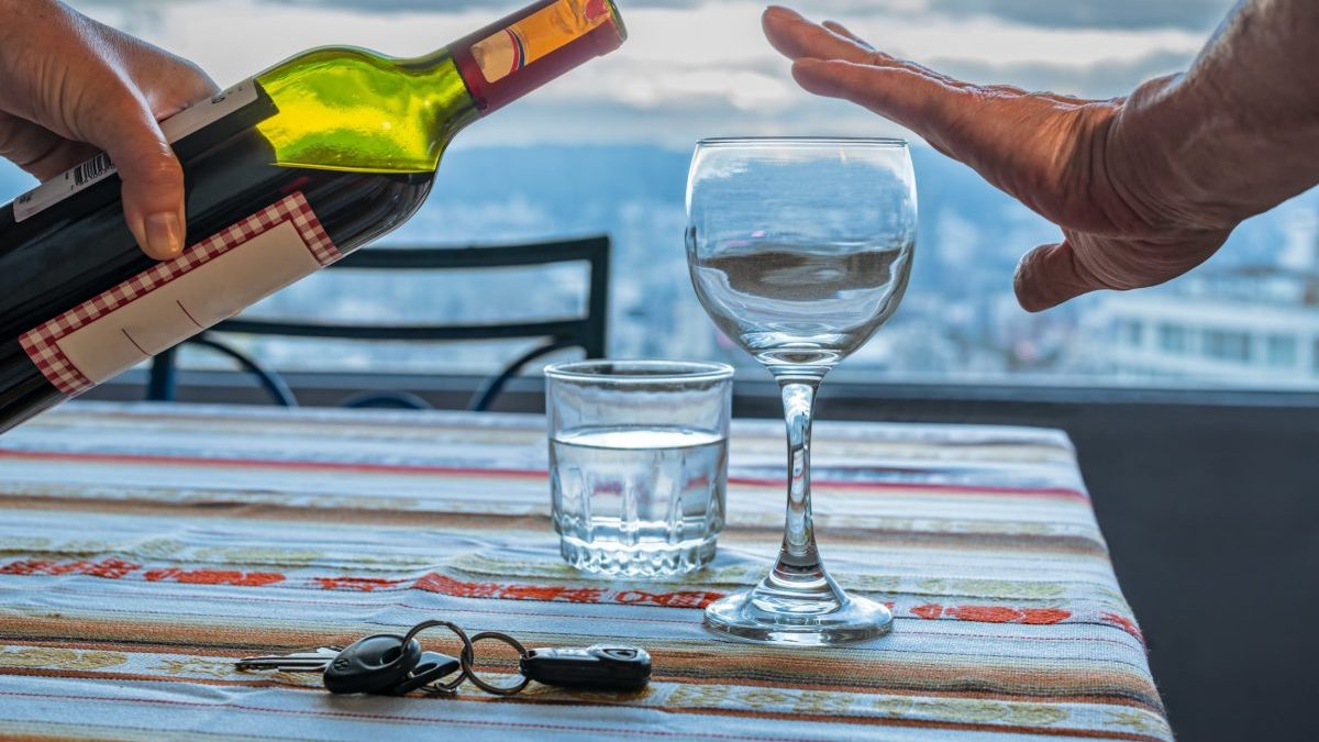 Close up of a hand of a man refusing red wine served with a bottle by a woman's hand, with car keys and a glass of water on a table at home.