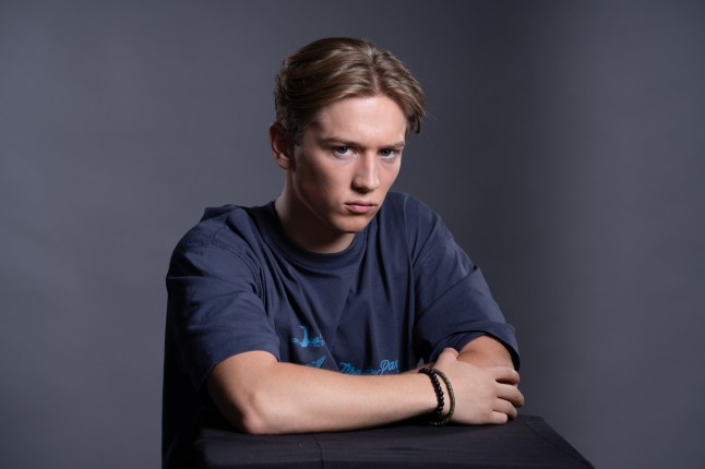 Max Murray posing as character Joel Marshall in an EastEnders promo. He's sitting on a chair, looking angrily into the camera, against a dark grey background.