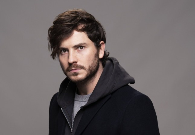 An EastEnders promo of Gray Atkins, looking seriously into the camera, and standing against a grey backdrop.