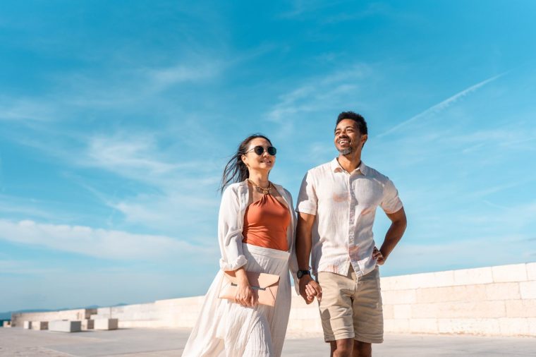 A mid-adult Black male and Asian female couple joyfully explore a charming coastal town under a clear blue sky. Their casual summer attire enhances the relaxed, adventurous atmosphere of Sutivan.
