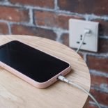 A mobile phone in a pink case charging battery by wire. A charging smartphone with cable on a wooden bedside table on a red brick wall backround