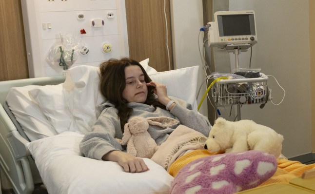 A police officer standing over Betsy Swain in a hospital bed and Dylan Wilson looking up at her from a chair in Corrie