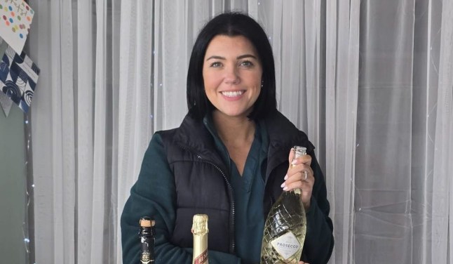 Natalie Dyson standing behind a table with champagne flutes, plastic cups, cupcakes and flowers, holding a bottle of champagne (Picture: Natalie Dyson)