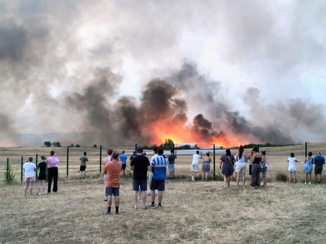 TOPSHOT - Bystanders look on as fire and smoke billow across Castres-Mazamet aerodrome, near Labruguiere in south-western France on July 24, 2026. France and Spain evacuated more than 160,000 people as unprecedented forest fires ravaged areas on July 25, 2026, near Madrid and Bordeaux, a French city flanked by tourist havens and world-renowned vineyards. French President Emmanuel Macron asked the army to mobilise to help contain the worst wildfire the country has ever seen after seeking European Union help, as a local official warned the blaze was heading towards Bordeaux. (Photo by Daniel ABELOUS / AFP via Getty Images)