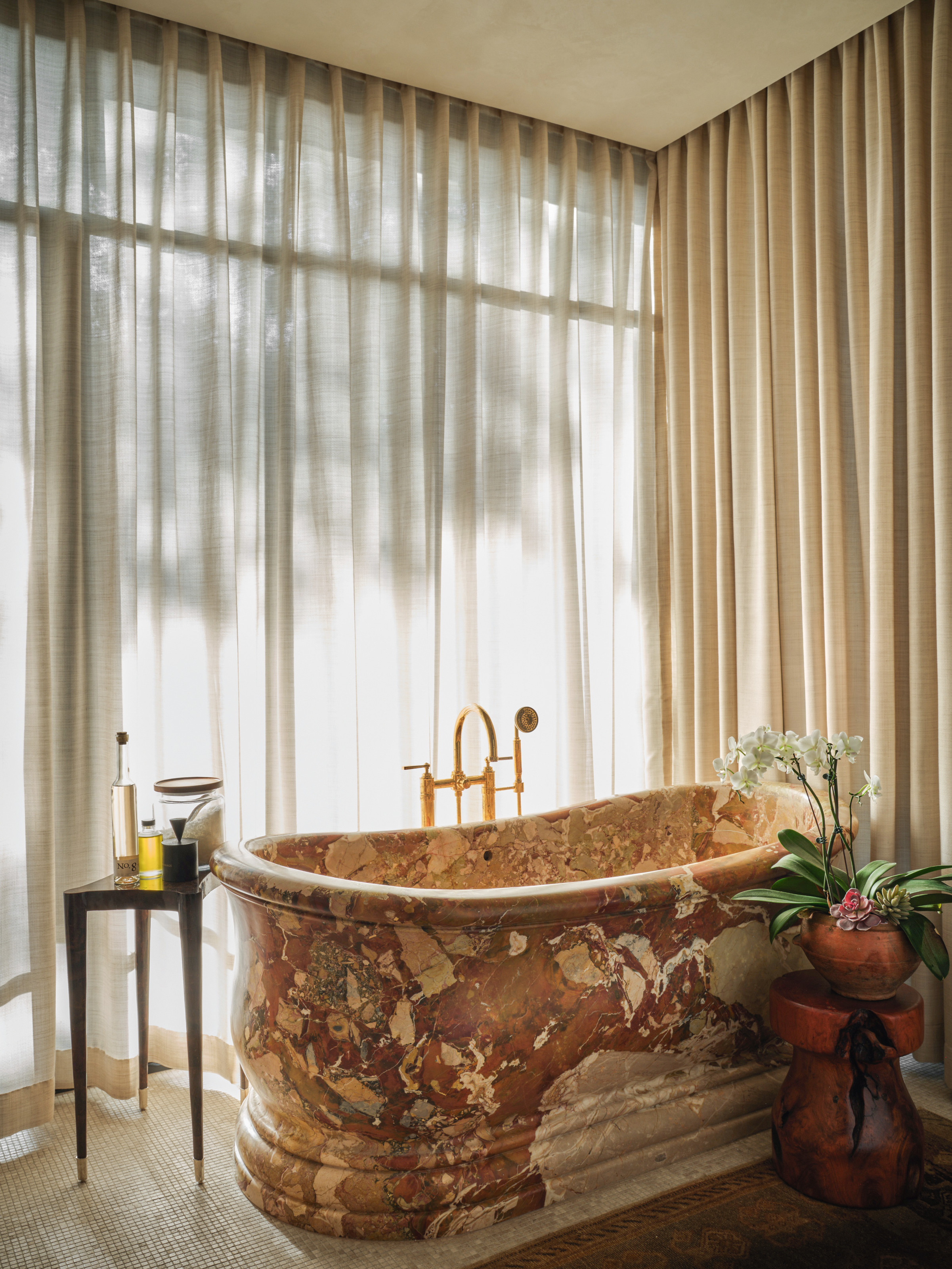 Beige bathroom with red and beige stone bath, black side table and terracotta stool