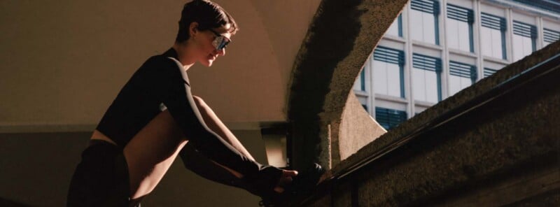 Person with short hair and sunglasses ties their shoe on a concrete ledge outdoors, with a building featuring blue window shades in the background.