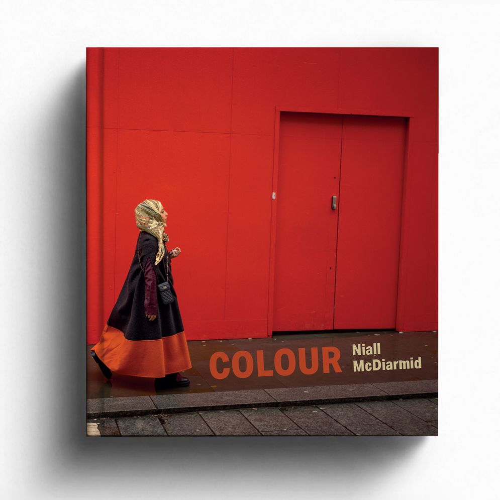 The front cover of a photography book titled "COLOUR" by Niall McDiarmid, featuring a person in a long dark coat with a patterned headscarf walking past a vibrant, solid red wall and doorway.