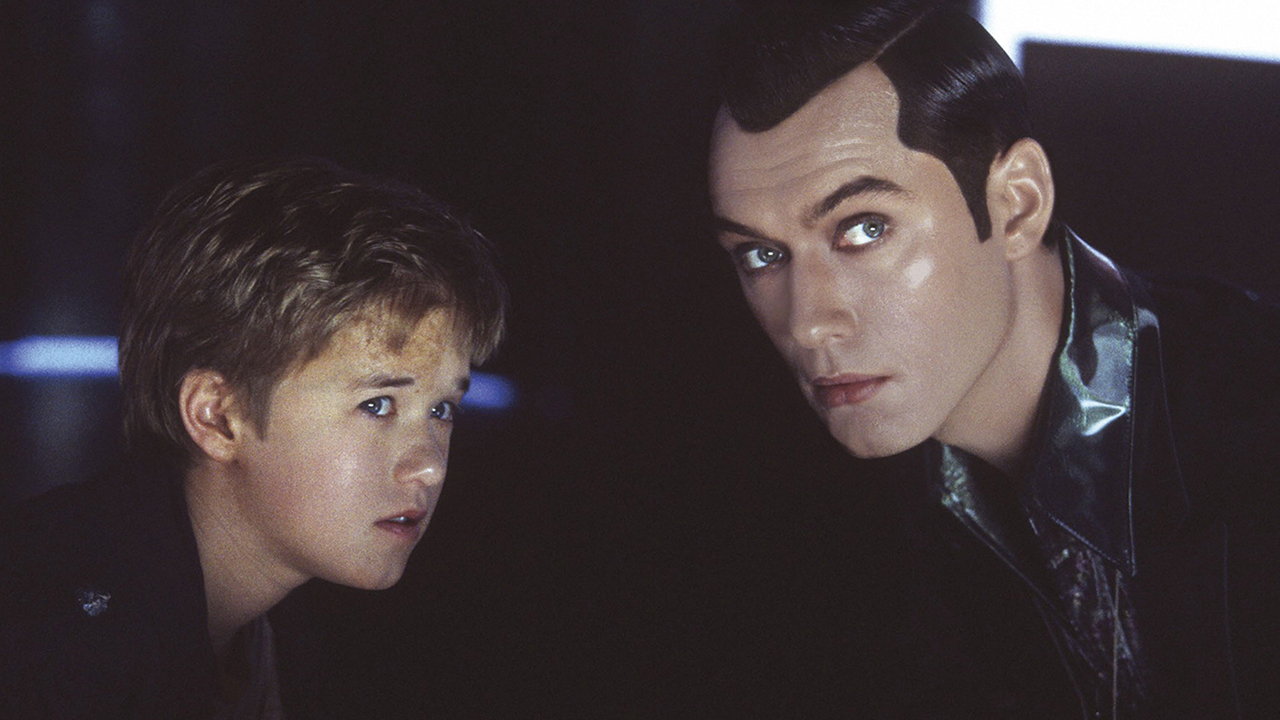 Still from the movie "A.I. Artificial Intelligence." Left is a young boy who is actually a robot. On the right is a man with slick black hair. They're both looking at something in the distance.