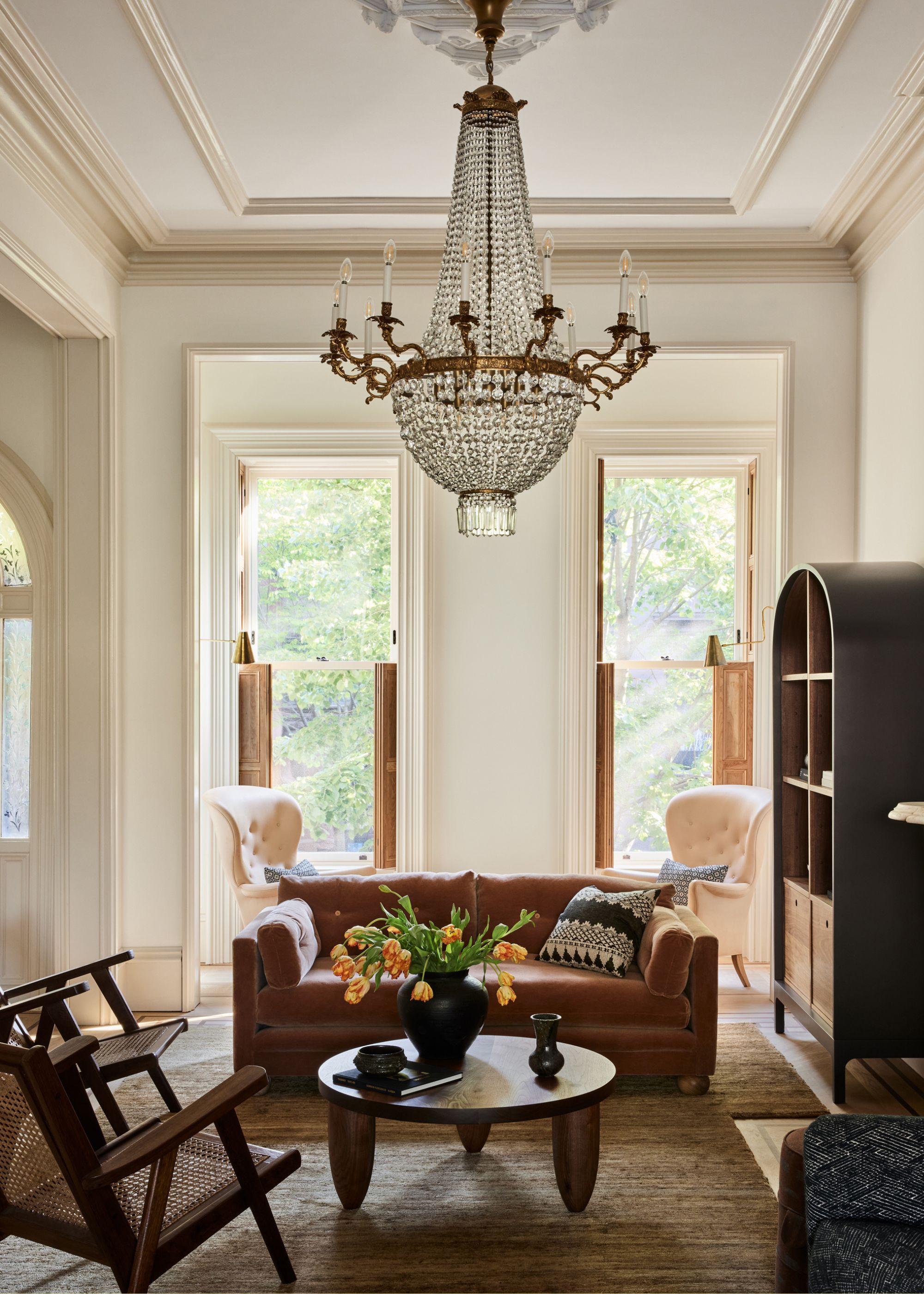 A bright living room with a crystal chandelier over a couch, rattan armchairs, a wood coffee table with a vase of tulips, and a pair of white armchairs