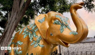 An elephant sculpture is pictured next to a tree. It is gold in colour with green leaves and foliage painted around its body.