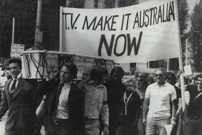 A black and white image of men carrying a coffin down a city street with a banner saying "TV make it Australian now".