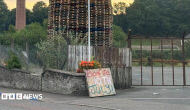 A bonfire of wooden pallets sits behind a fence, in front of the fence is a brick wall and a wooden sign is propped up against it with "bonfire 10th July" written on it